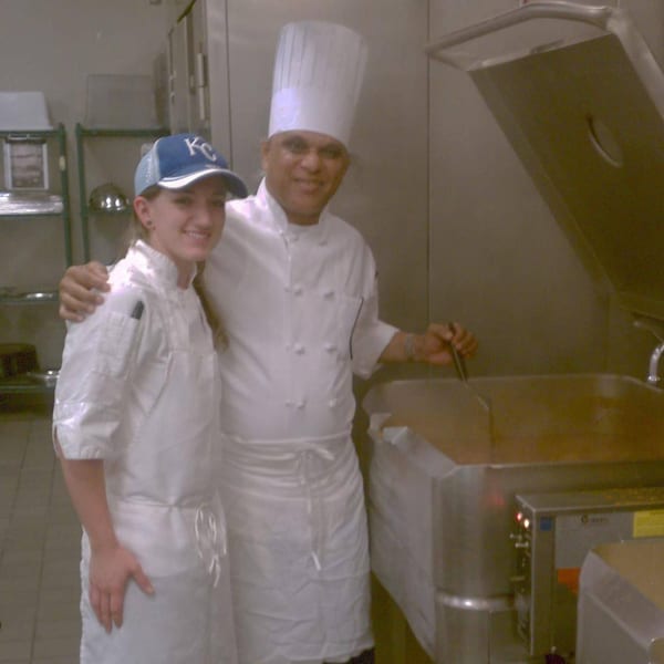 Two chefs, one in a tall white hat, smiling in a professional kitchen with large cooking equipment.