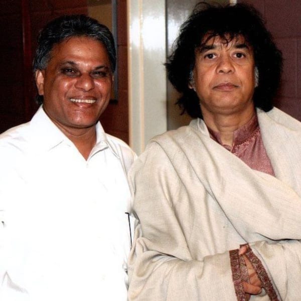 Two men, one smiling in a white shirt and another in a shawl, pose together.