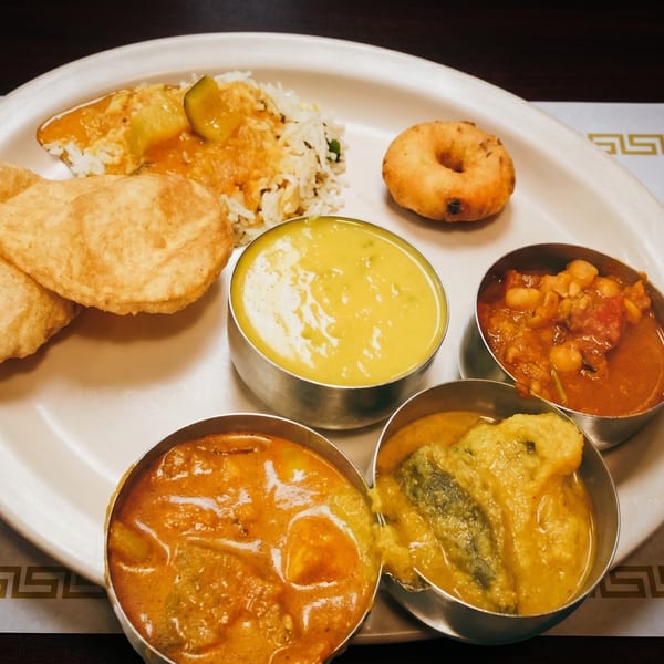 A white plate with various Indian dishes, including rice, curries, fried bread, and a vada.