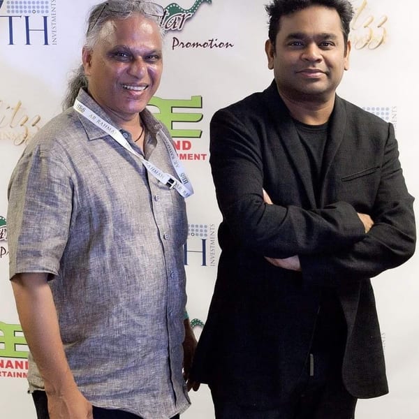 Two men, one with long gray hair and a gray shirt, the other in a black suit, smiling.