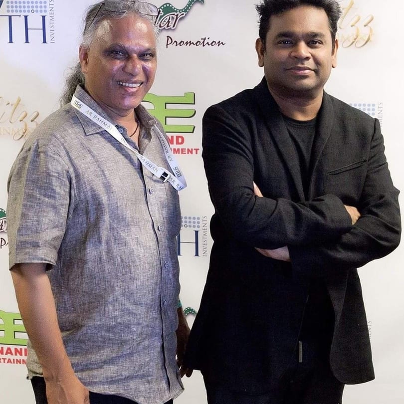 Two men, one with long gray hair and a gray shirt, the other in a black suit, smiling.