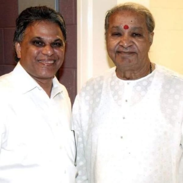 Two smiling men in white shirts, one with a red bindi, stand side-by-side.