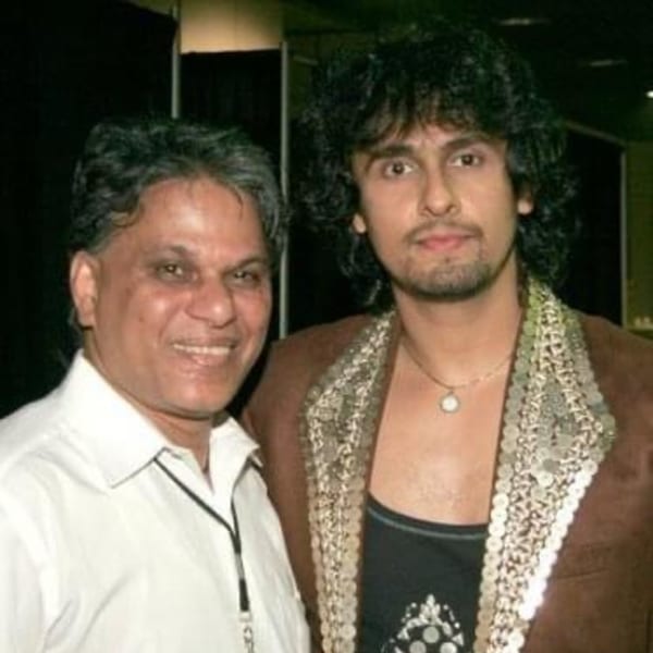 Two men, one older with a white shirt and one younger with curly hair and a brown jacket.