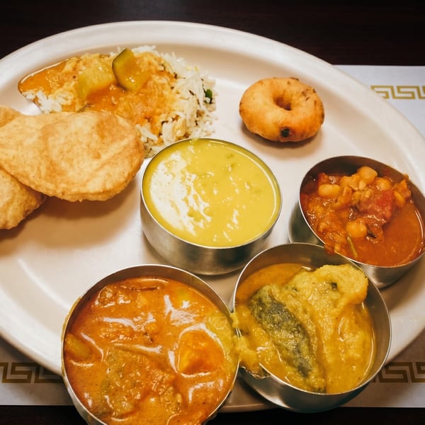 A white plate holds an Indian thali meal with rice, puri, vada, and four bowls of curries.