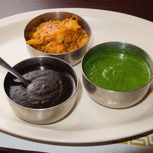 Three metal bowls of Indian chutneys: orange potato, dark brown, and bright green, on a white plate.
