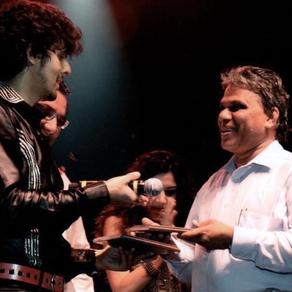 Two men on stage, one holding a microphone, the other receiving awards, with a woman in the background.