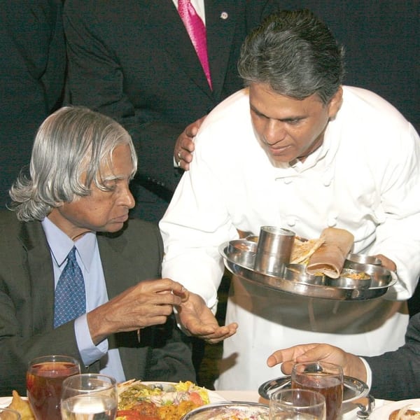 A chef in a white uniform serves a meal on a silver platter to a seated man in a suit.