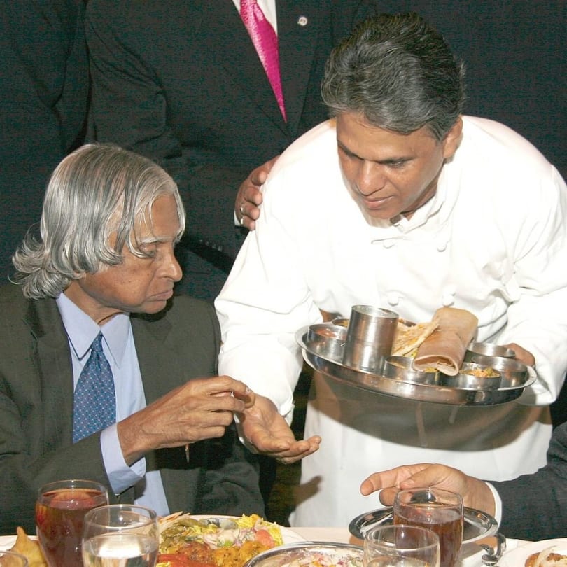 A chef in a white uniform serves a meal on a silver platter to a seated man in a suit.