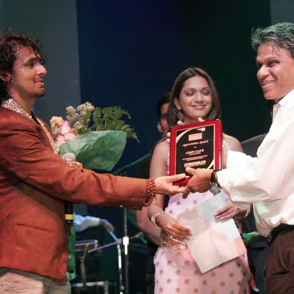 A man in a brown jacket receives a bouquet and an award plaque from another man.