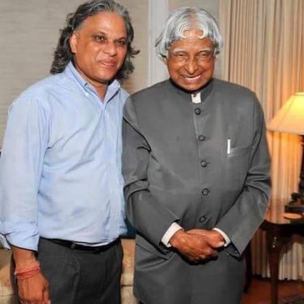 Two men, one with long dark hair and a light blue shirt, the other in a dark suit, smiling.