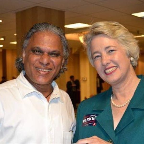 A man in a white shirt and a woman in a teal jacket with a pearl necklace smile.