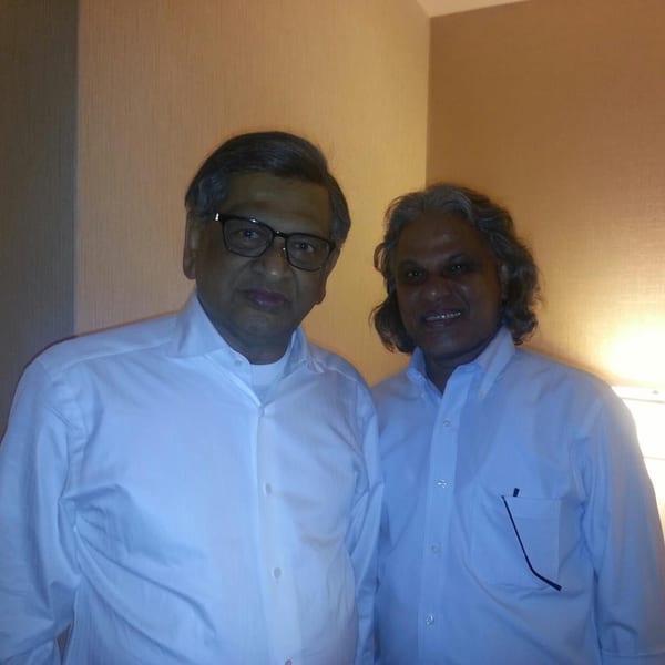 Two men in light blue button-down shirts stand side-by-side, smiling at the camera.