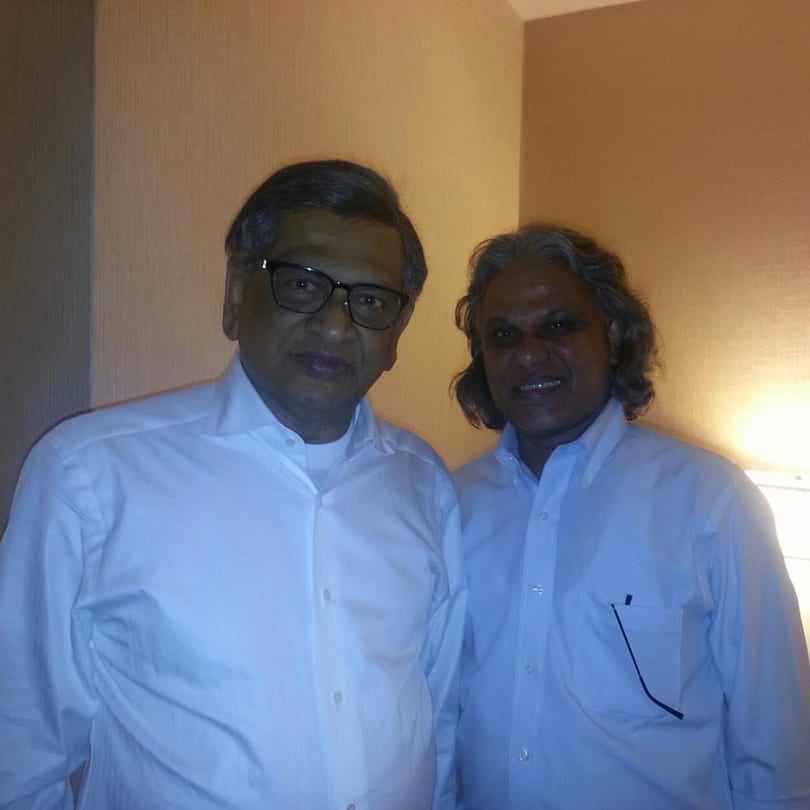 Two men in light blue button-down shirts stand side-by-side, smiling at the camera.
