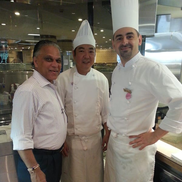 Three men, two in chef's whites and one in a plaid shirt, smiling in a professional kitchen.