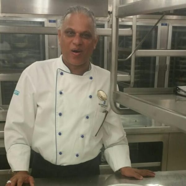 A male chef in a white uniform with blue buttons stands in a commercial kitchen.