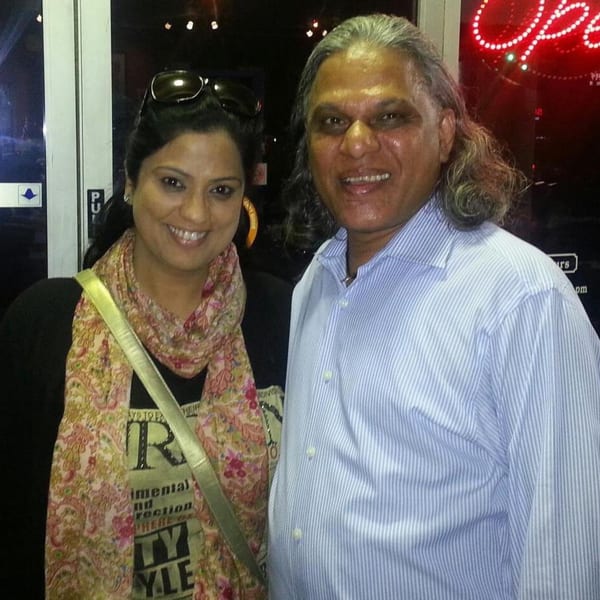 A man and woman smiling, standing together in front of a neon "Open" sign.