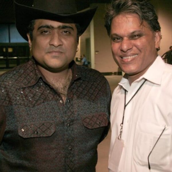 Two men, one in a cowboy hat and patterned shirt, the other in a white shirt, smiling at the camera.