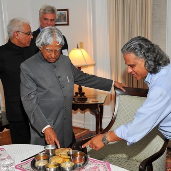 Four men in a room, two pointing at a tray of Indian food on a table.
