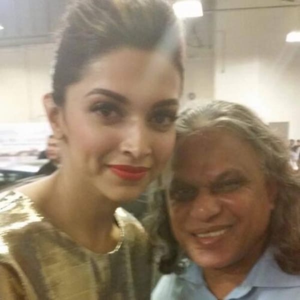 Deepika Padukone, in a gold top, smiles next to a man with long gray hair in a blue shirt.