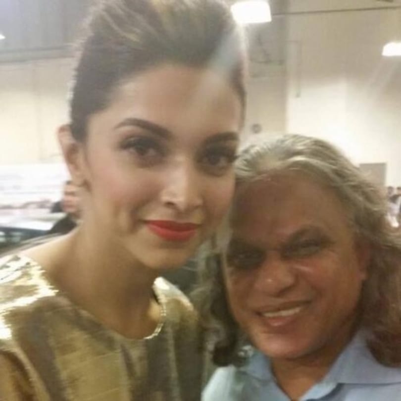 Deepika Padukone, in a gold top, smiles next to a man with long gray hair in a blue shirt.