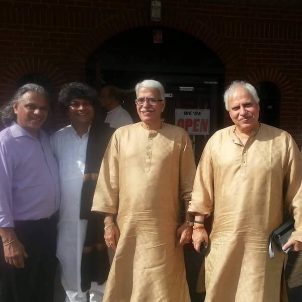 Four men, two in traditional Indian attire, stand smiling in front of a brick building.