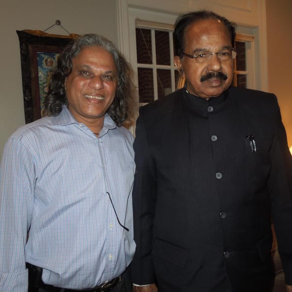 Two men, one in a blue plaid shirt and the other in a black suit, stand side-by-side.