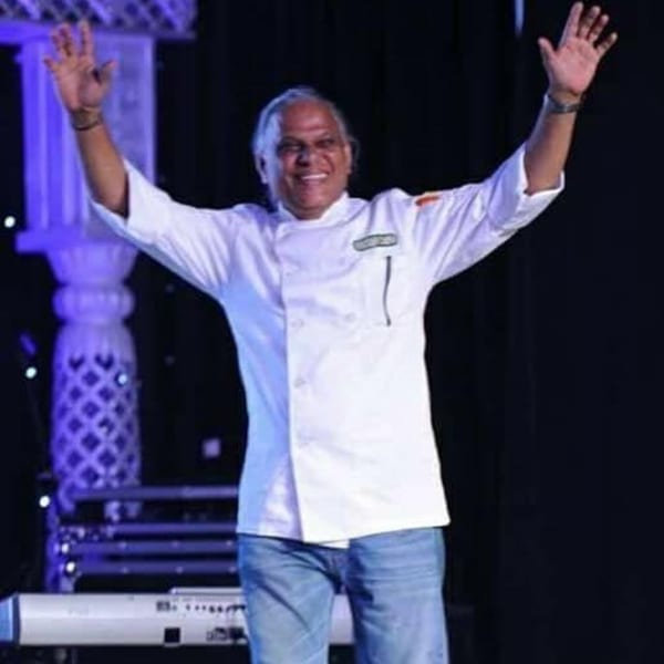Smiling chef in a white jacket and jeans, arms raised in celebration on a stage.