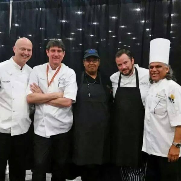 Five chefs, three in white coats and two in black aprons, smiling in front of a dark curtain.