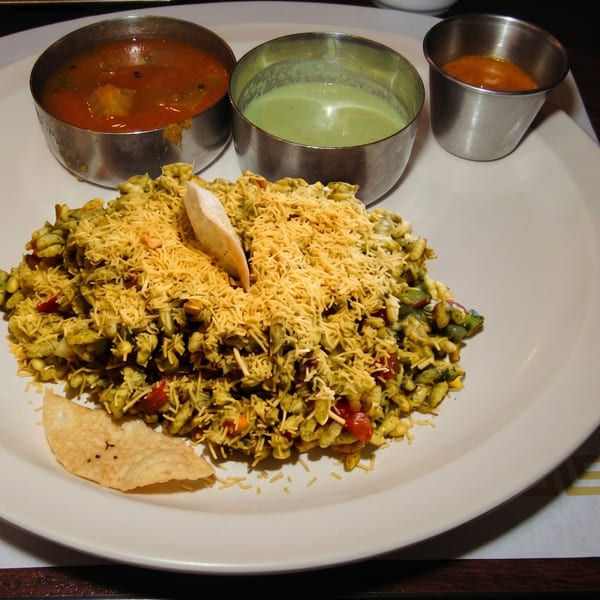 A plate of Indian Bhel Puri with three small metal bowls of chutneys.