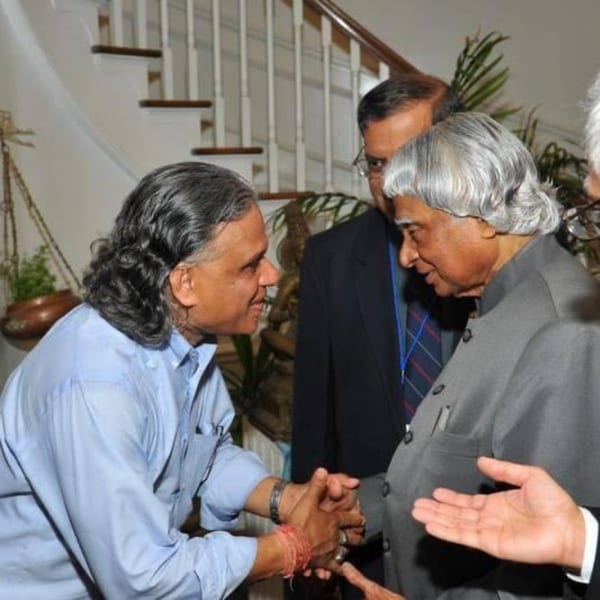 Two men, one with long dark hair and one with white hair, shake hands indoors.