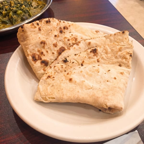 Three pieces of golden-brown flatbread with charred spots on a white plate, with a curry dish in the background.