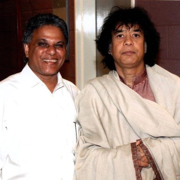 Two men, one smiling in a white shirt, the other in a shawl, pose together.