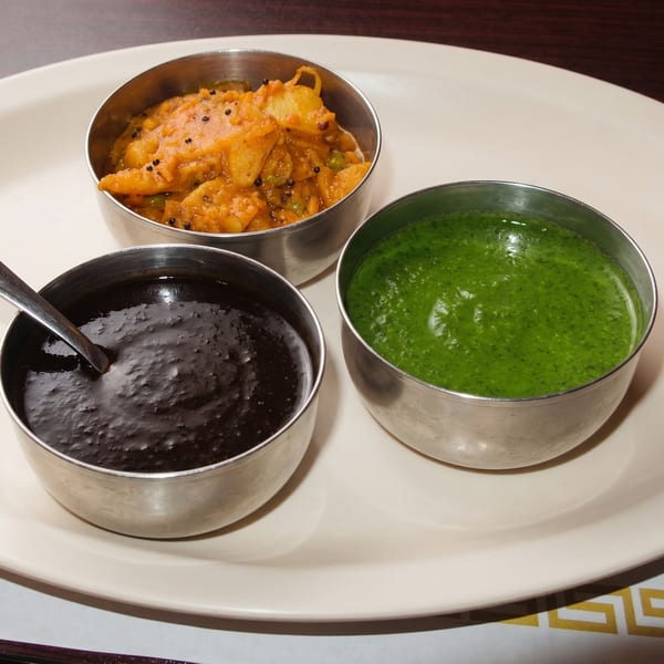 Three small metal bowls of Indian chutneys: orange, dark brown, and bright green, on a white plate.